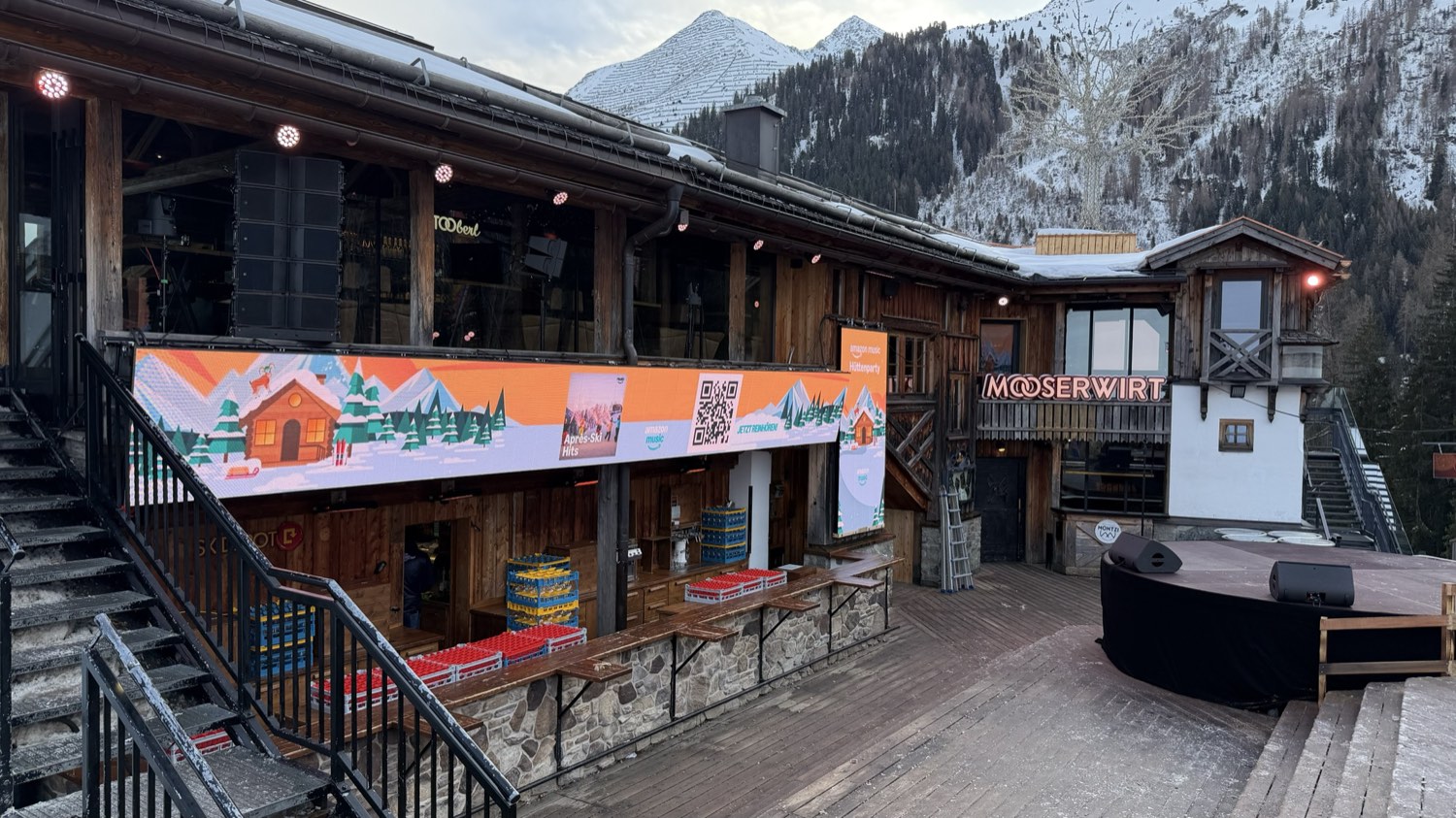 Große LED-Werbewand an der Fassade einer Skihütte, darüber Lautsprecheranlage und Scheinwerfer, rechts daneben eine Bühne, im Hintergrund verschneite Berge