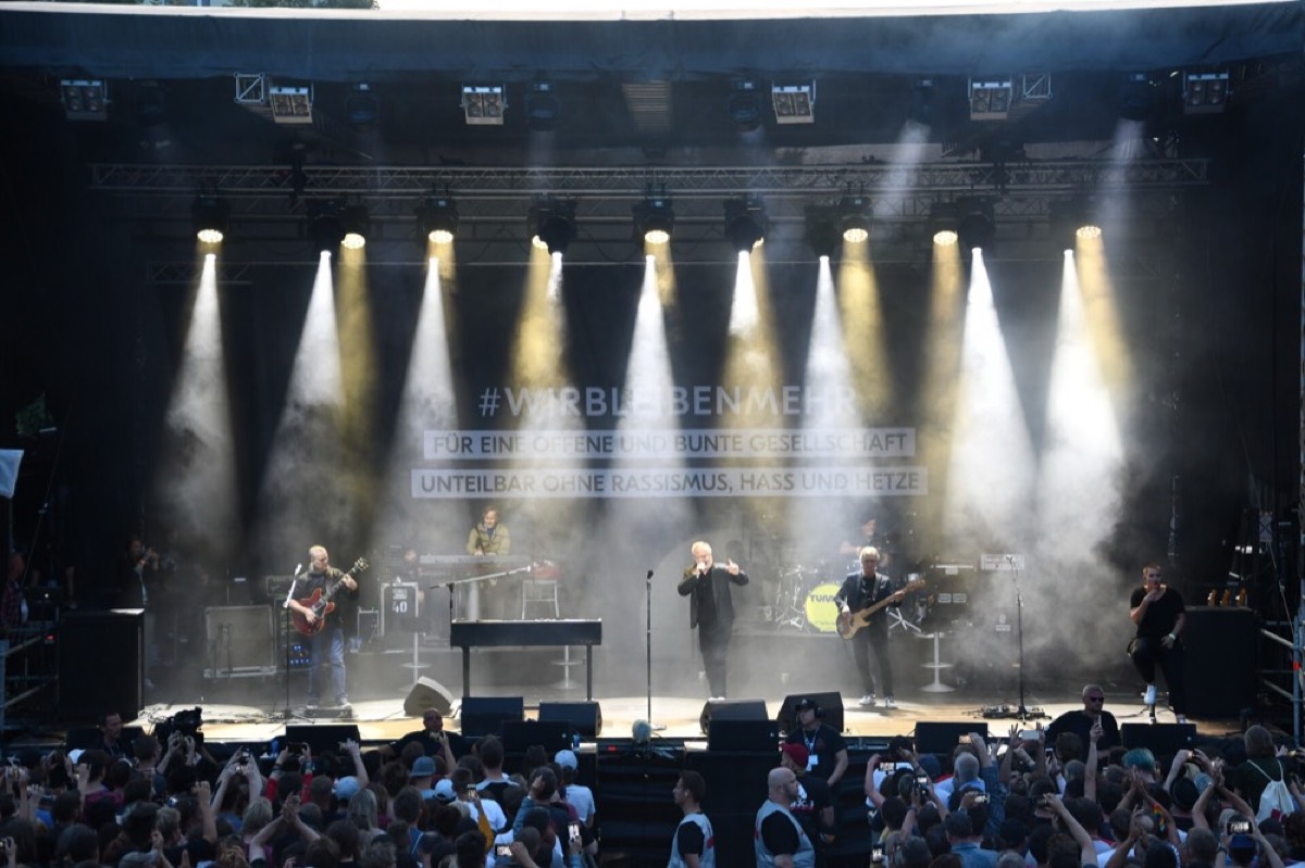 Konzertbühne mit Band, weiße Lichtstrahlen fallen durch Nebel auf die Bühne, davor das Publikum